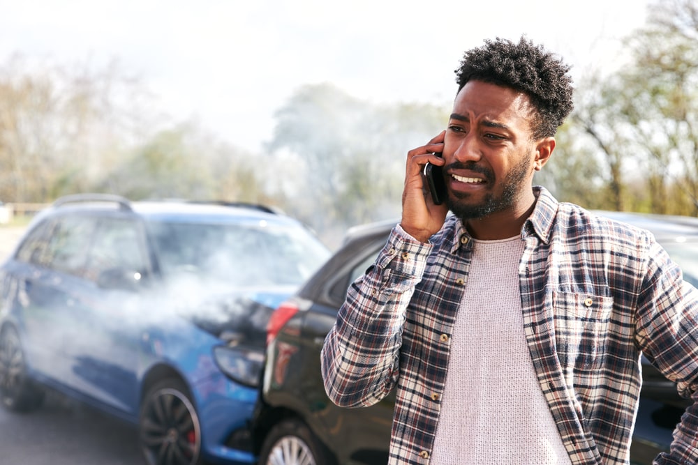 Young man standing by a damaged car after a road accident, reporting the incident to an insurance company using a mobile phone.